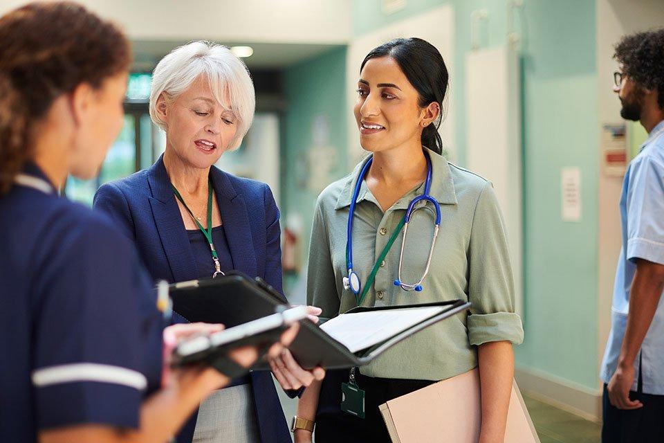 Three healthcare providers (a nurse, doctor, and healthcare administrator) discussing work in a hospital hallway.